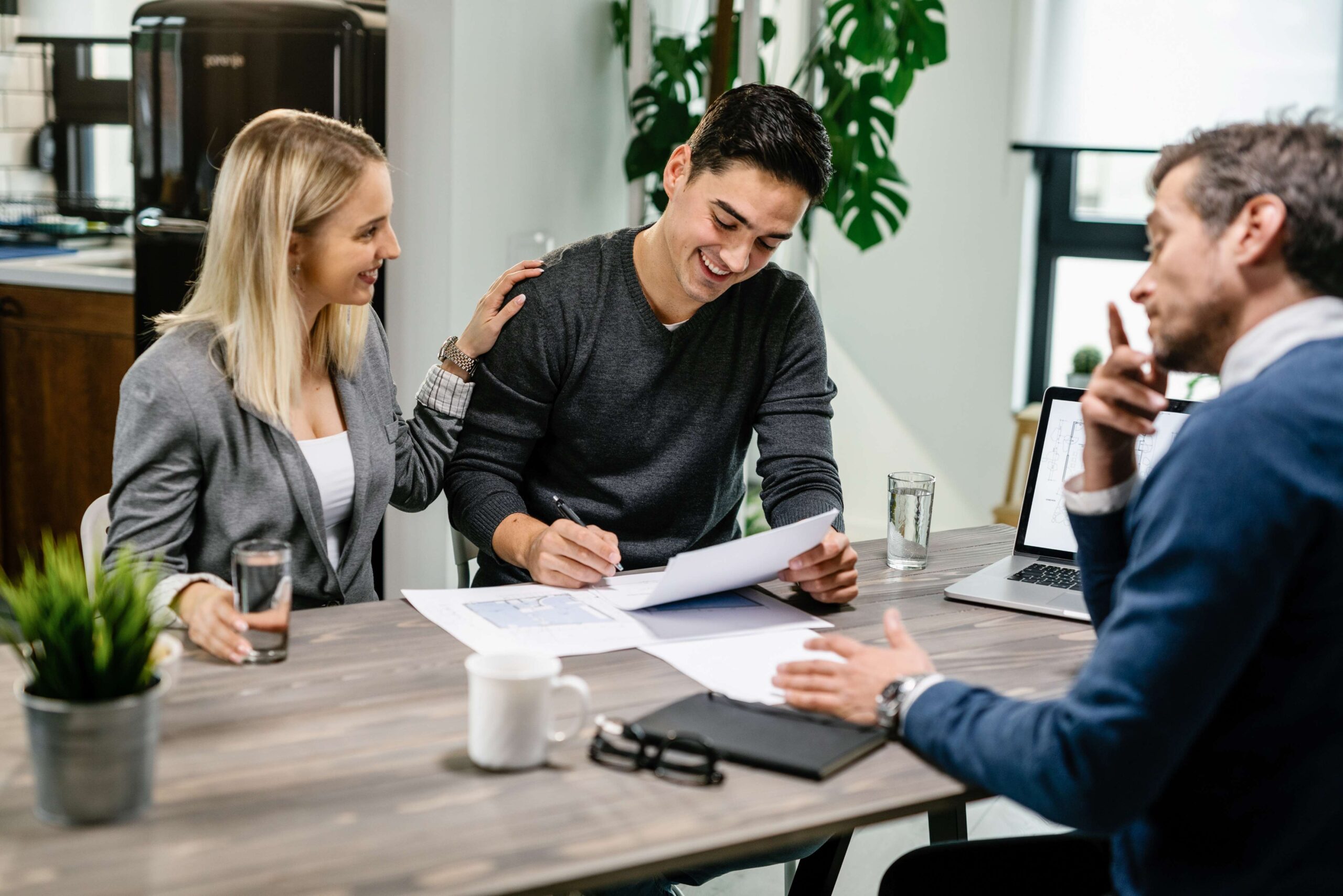 Joven hombre feliz firmando un contrato hipotecario mientras está con su esposa en una reunión con un agente de bienes raíces