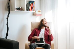 Joven sonriente en el interior de su casa disfrutando de su espacio preferido dedicado a la comodidad y la lectura iluminado por la iluminación natural que pasa a través de los ventanales