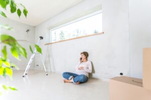 Mujer joven usando el teléfono mientras está sentada y recargada en la pared blanca que genera un ambiente más brillante en la habitación